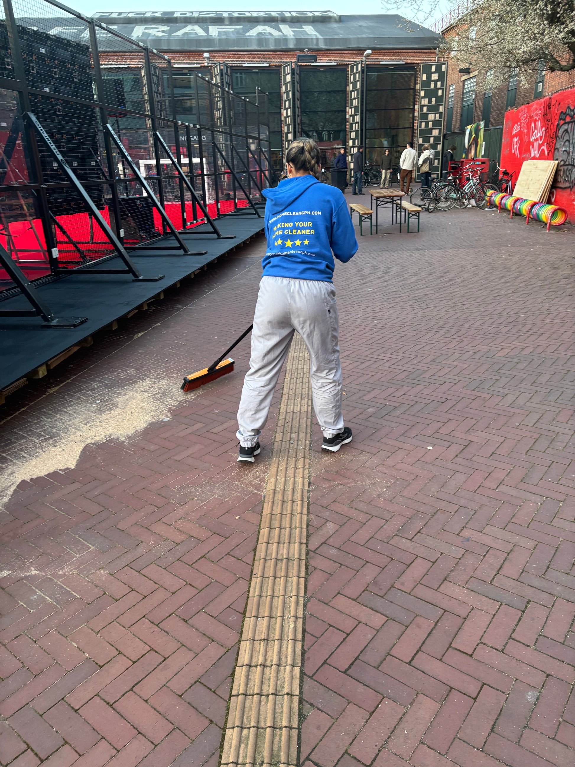 Person cleaning outdoor event space with a broom in Copenhagen, wearing a blue jacket and gray sweatpants.