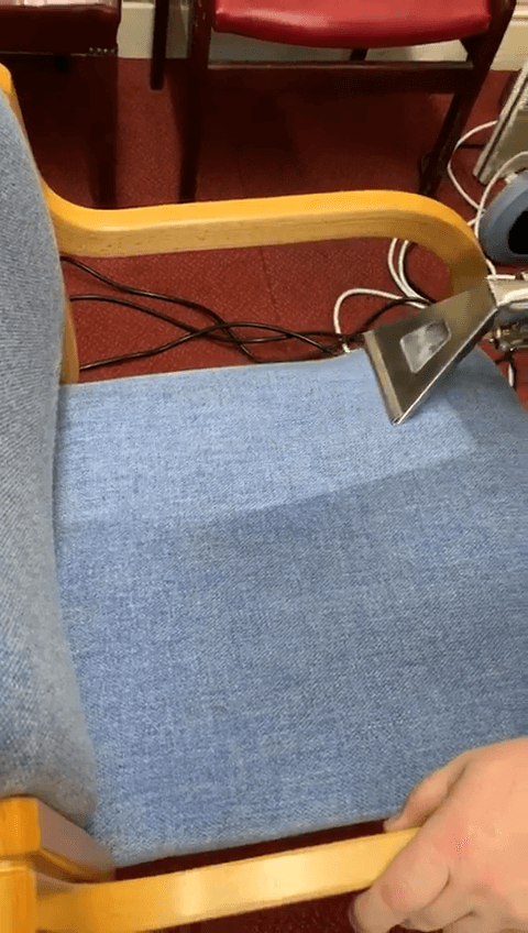 Close-up of a hand using a cleaner on a blue upholstered chair in a cleaning process.